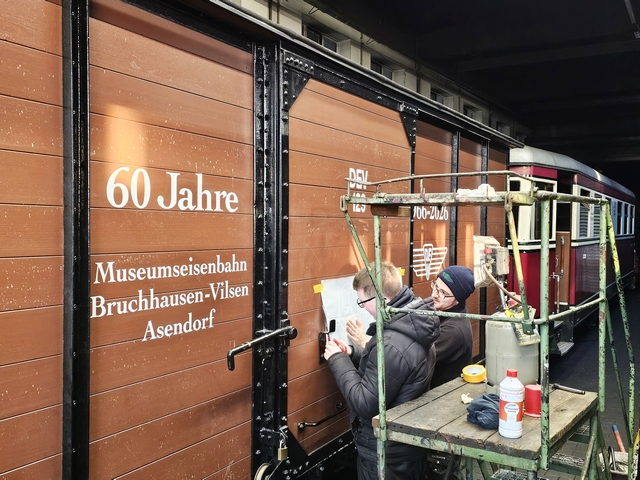 Abb.: Jonas Kindermann und Eike Amelung beim Aufbringen der Beklebung in der DEV-Werkstatt. Das Wetter spielte nicht ganz mit, zur Sicherstellung der Verklebung war daher der rechts stehende Gas-Heizstrahler erforderlich (Klicken vergrößert).